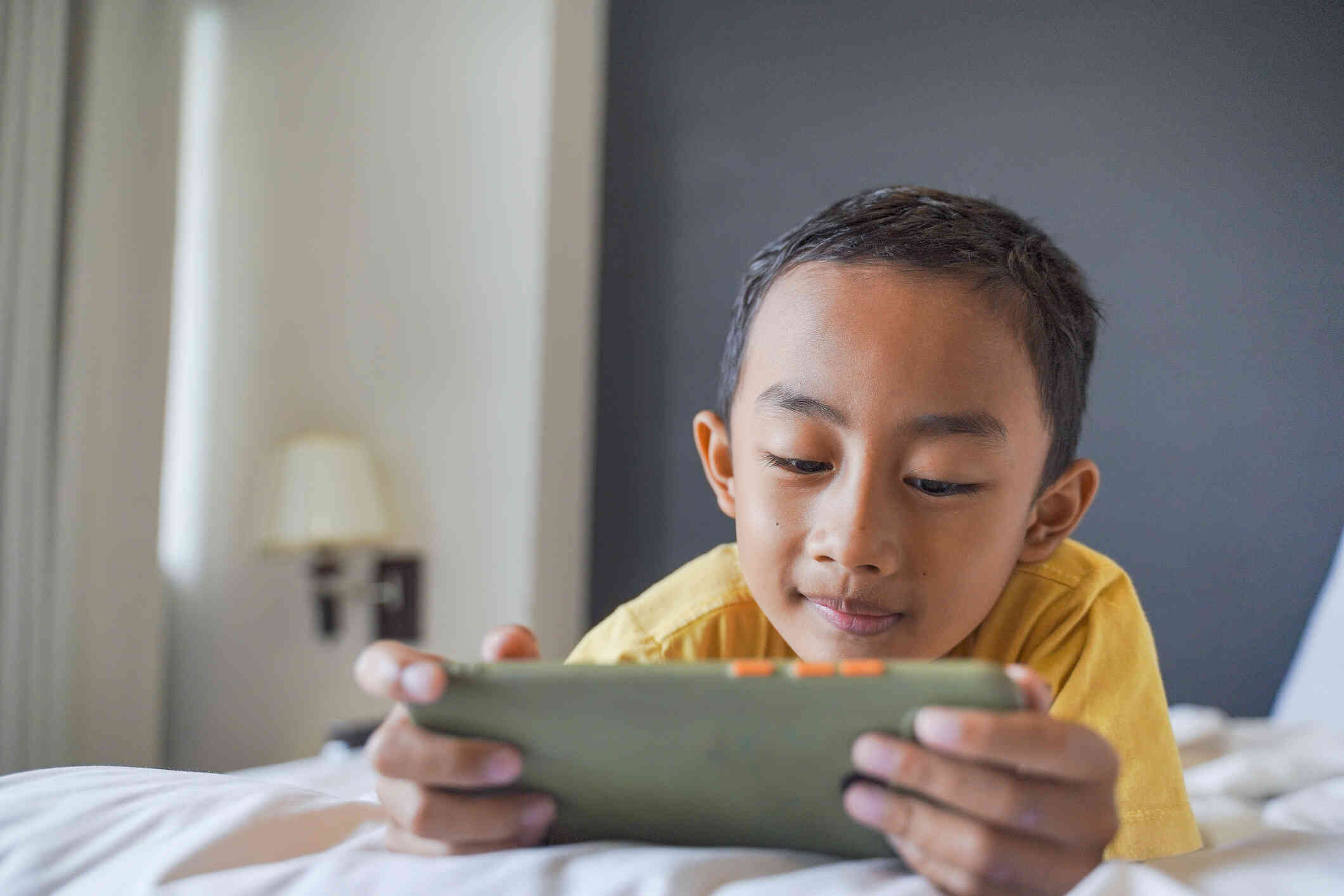 A young boy lays on his stomach on the bed and looks at the phone in her hand.